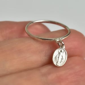 Peut inclure: Une bague en argent avec un petit charme de forme ovale suspendu. Le charme repr&eacute;sente une figure religieuse.