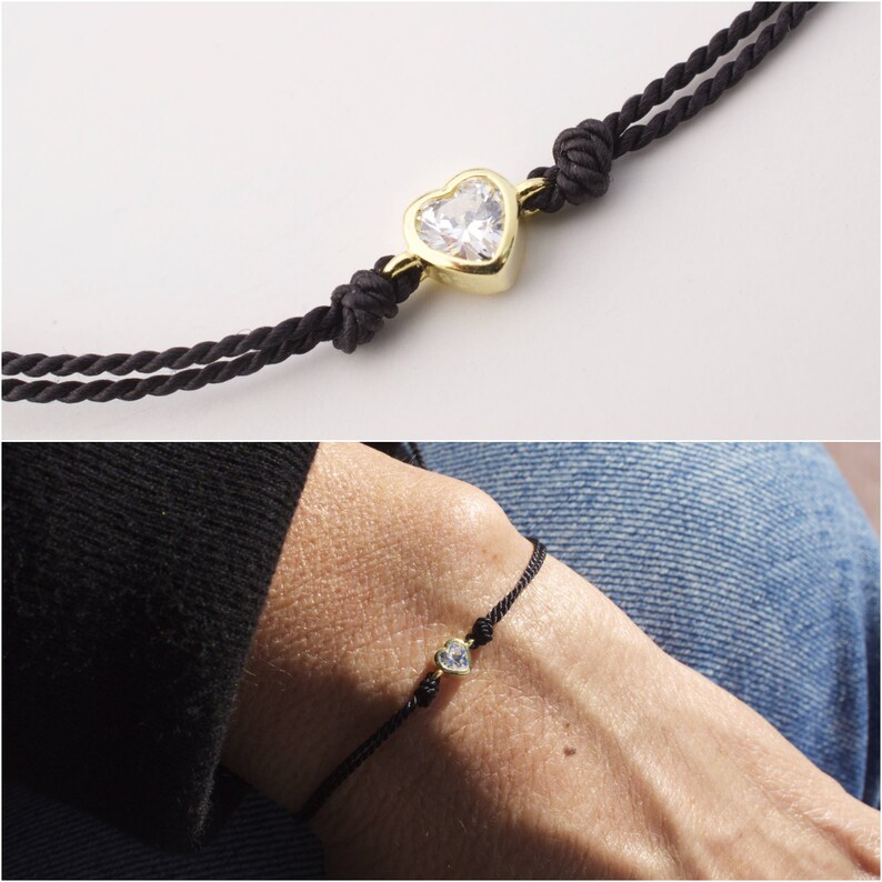 May include: A black cord bracelet with a gold heart-shaped charm featuring a clear crystal. The bracelet is shown on a person's wrist.