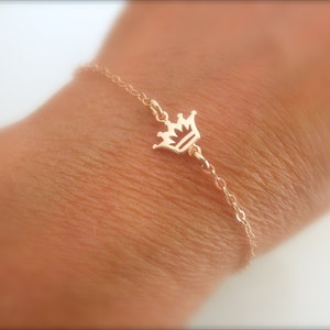 May include: A delicate gold-colored bracelet featuring a small crown charm. The crown is centered on a fine chain, creating a simple yet elegant piece of jewelry. The bracelet is designed to be worn on the wrist.