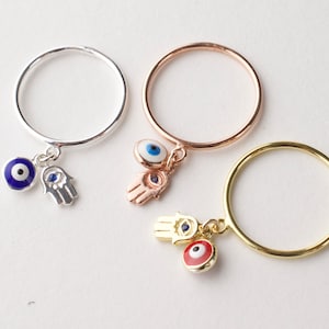 May include: Three rings in silver, rose gold, and gold. Each ring features a charm with an evil eye and a Hamsa hand. The evil eye charms are blue, white, and red. The Hamsa hand charms are silver, rose gold, and gold.