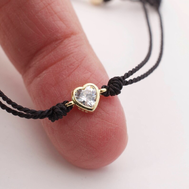 May include: A black string bracelet with a gold heart-shaped charm featuring a clear crystal.