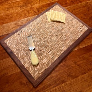 May include: A wooden cheese board with a geometric cube pattern, a dark brown border, and a light-colored handle cheese knife. Three crackers are placed on the board. The board is on a wooden surface.