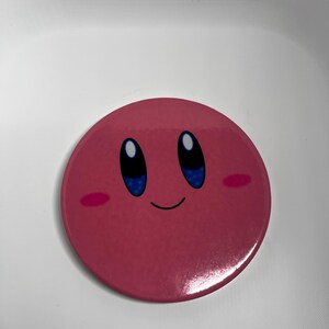 May include: A circular, pink badge featuring the character Kirby. The badge has a glossy finish and depicts Kirby's face with blue eyes, a small smile, and pink cheeks. The background is white.
