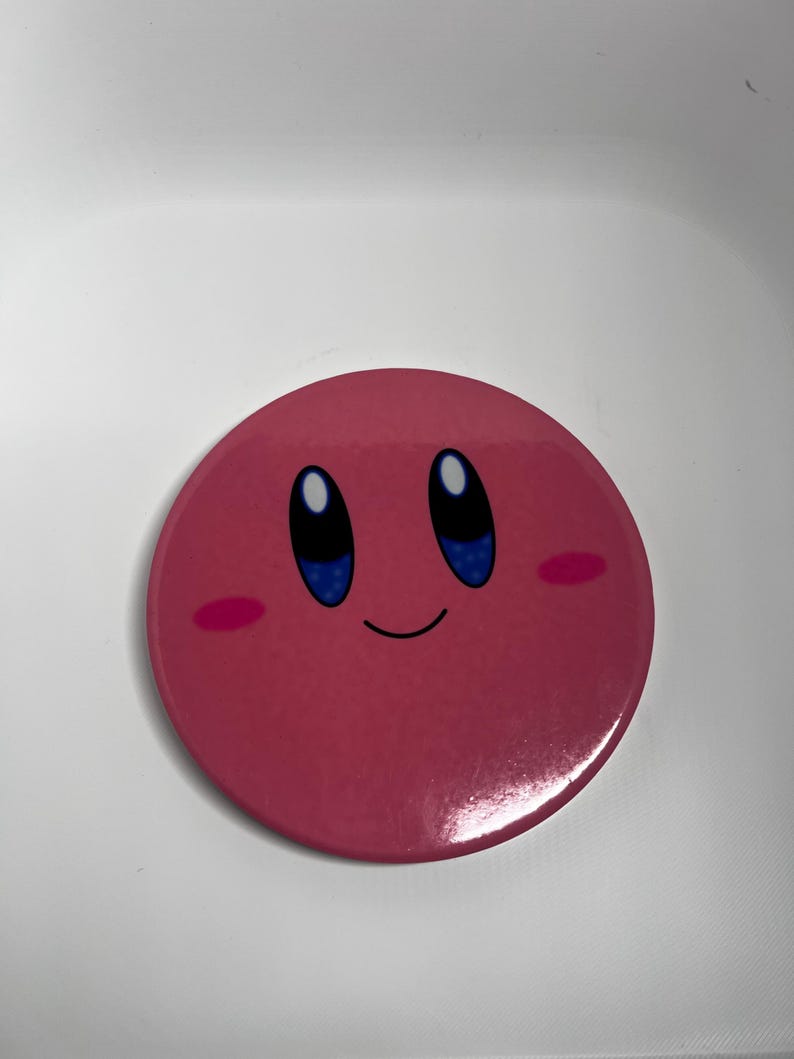 May include: A circular, pink badge featuring the character Kirby. The badge has a glossy finish and depicts Kirby's face with blue eyes, a small smile, and pink cheeks. The background is white.