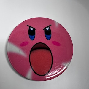 May include: A circular pink badge with a cartoon character's face. The character has an open mouth, blue eyes, and angry eyebrows. The badge has white streaks and a red tongue. The design is on a white background.