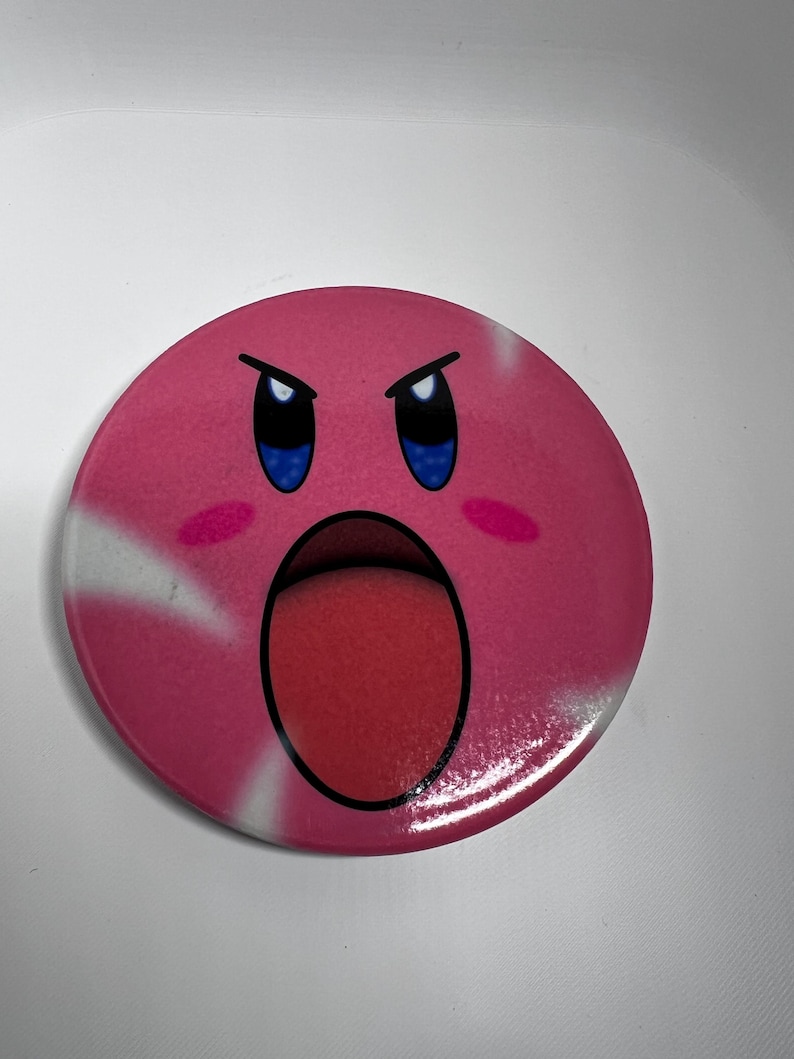 May include: A circular pink badge with a cartoon character's face. The character has an open mouth, blue eyes, and angry eyebrows. The badge has white streaks and a red tongue. The design is on a white background.