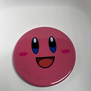 May include: A circular, pink badge displaying a smiling cartoon character with blue eyes, a red mouth, and pink cheeks. The badge has a shiny surface and is set against a white backdrop. The character's design is simple and easily recognised.