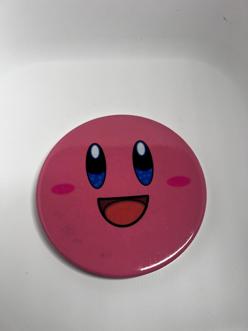 May include: A circular, pink badge displaying a smiling cartoon character with blue eyes, a red mouth, and pink cheeks. The badge has a shiny surface and is set against a white backdrop. The character's design is simple and easily recognised.