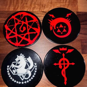 May include: A set of four black, round coasters with red graphic designs. Each coaster displays a different design: a pentagram, a dragon, a horse, and a symbol with a crown and wings. The coasters are made of a glossy material.