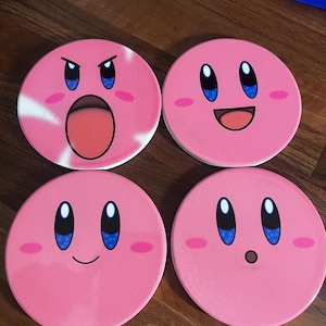 May include: Four pink, circular coasters featuring the character Kirby with various facial expressions. Each coaster has a white border and blue eyes. One Kirby has an open mouth, another is smiling, and two others have neutral expressions.