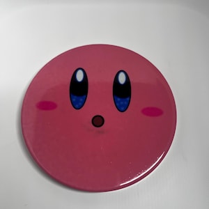 May include: A circular, pink button featuring the character Kirby with large blue eyes, rosy cheeks, and a small, open mouth. The button has a glossy finish against a white background.