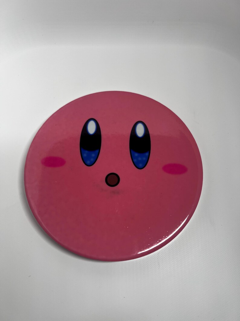 May include: A circular, pink button featuring the character Kirby with large blue eyes, rosy cheeks, and a small, open mouth. The button has a glossy finish against a white background.