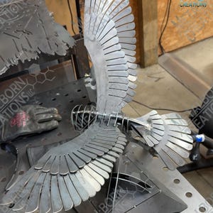 May include: A metal sculpture of a bird with wings spread wide, constructed from individual silver-coloured steel plates welded together. The bird features a wire-frame head and a layered feather design on its wings and tail, mounted on a base.