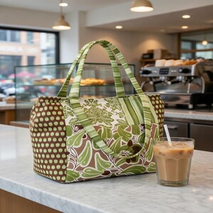 Puede incluir: Un bolso tote rectangular con dise&ntilde;o de patchwork que presenta patrones circulares en marr&oacute;n y verde, un estampado de damasco blanco y verde en el centro y motivos de hojas verdes. El bolso tiene dos asas de tela a rayas.