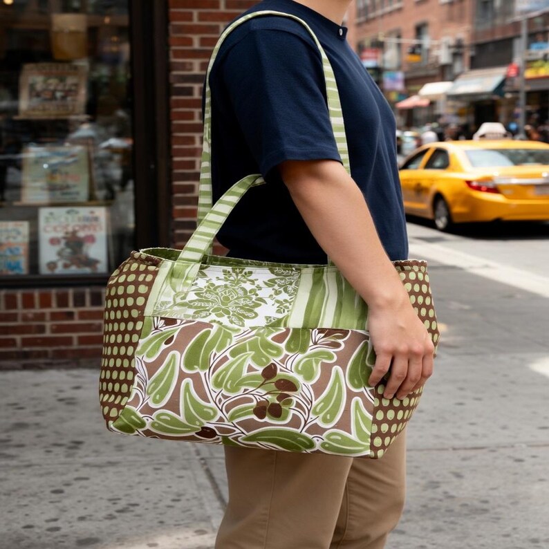 Puede incluir: Un bolso tote con dise&ntilde;o de patchwork que presenta patrones de hojas verde oliva, un panel floral blanco y paneles laterales marrones con un patr&oacute;n de puntos peque&ntilde;os. El bolso tiene dos asas a rayas a juego y forma rectangular.