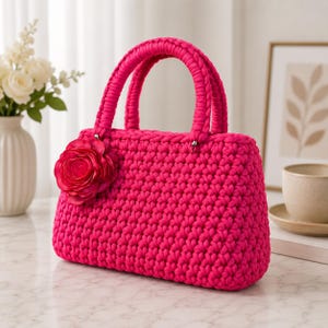May include: A vibrant fuchsia handbag crafted from thick, chunky yarn in a dense crochet stitch. The bag features two rounded top handles and a large, layered fabric flower embellishment in a matching shade of pink attached to the front side.