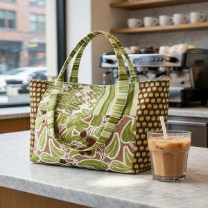 Puede incluir: Un bolso de tela con dise&ntilde;o de patchwork en tonos verdes y marrones, con patrones de hojas, rayas y puntos. El bolso tiene dos asas de tela a juego y una base rectangular. Junto a &eacute;l hay un vaso de caf&eacute; helado con una pajita blanca.