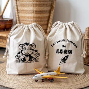 May include: Two beige drawstring bags with black graphics. One bag features a monkey family illustration, the other has text that reads "Les petits bidules de ADAM" with a rocket ship graphic. A yellow and white toy airplane sits in front.