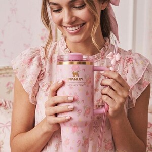 K&ouml;nnte beinhalten: Ein rosa Stanley-Becher mit Blumenmuster, Griff und klarem Strohhalm. Auf dem Becher steht "Stanley" und "LOVESHACKFANCY". Eine Person in einem rosa Blumenkleid h&auml;lt den Becher.