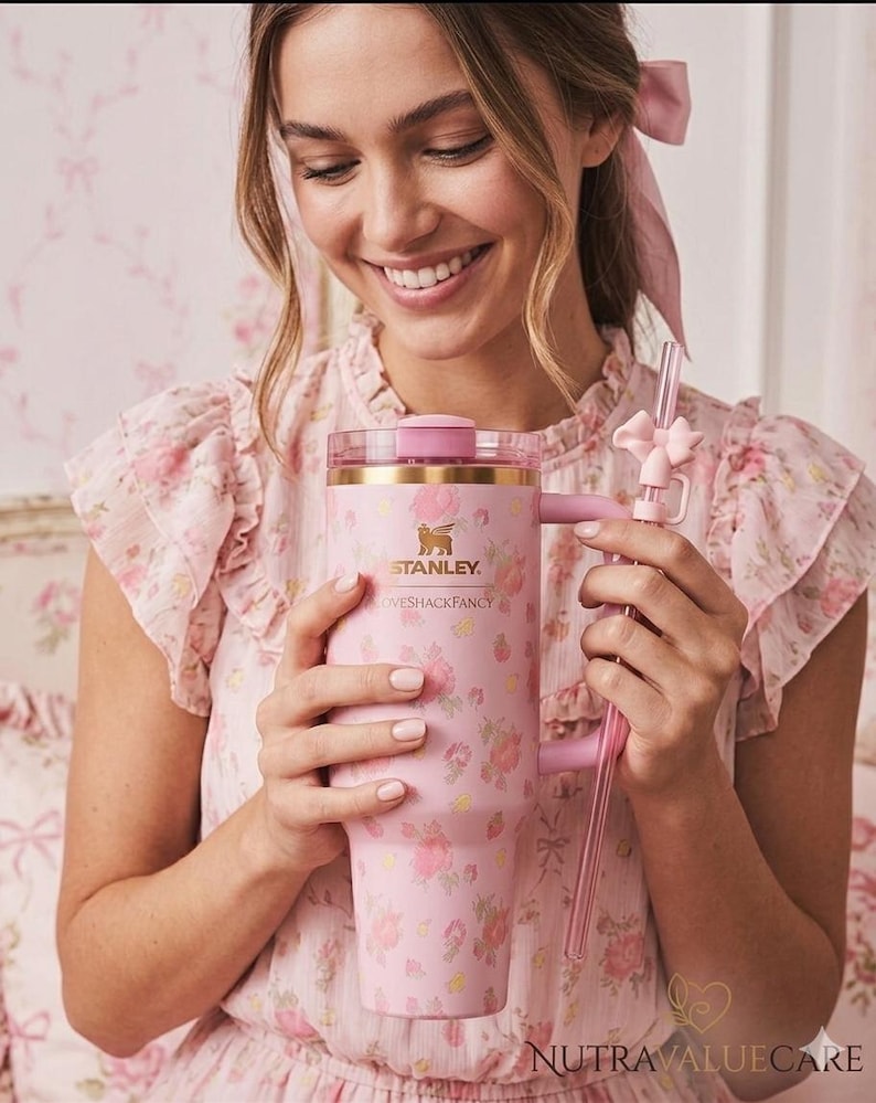 K&ouml;nnte beinhalten: Ein rosa Stanley-Becher mit Blumenmuster, Griff und klarem Strohhalm. Auf dem Becher steht "Stanley" und "LOVESHACKFANCY". Eine Person in einem rosa Blumenkleid h&auml;lt den Becher.
