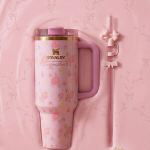 K&ouml;nnte beinhalten: Ein rosa Stanley Loveshackfancy-Becher mit floralem Muster, Griff und passendem Strohhalm mit Schleifenakzenten. Der Becher hat einen goldenen Rand und einen rosa Deckel. Das Set wird in einem rosa geformten Beh&auml;lter pr&auml;sentiert.