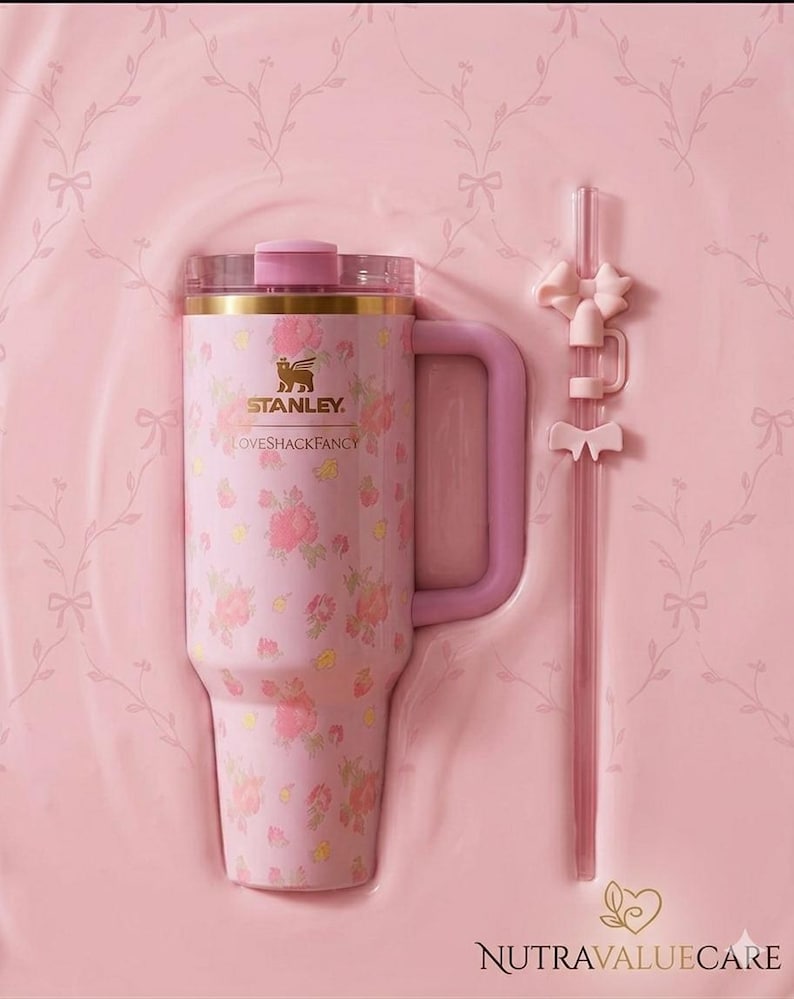 K&ouml;nnte beinhalten: Ein rosa Stanley Loveshackfancy-Becher mit floralem Muster, Griff und passendem Strohhalm mit Schleifenakzenten. Der Becher hat einen goldenen Rand und einen rosa Deckel. Das Set wird in einem rosa geformten Beh&auml;lter pr&auml;sentiert.