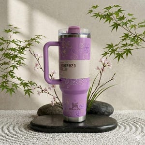 May include: A lavender Stanley Quencher H2.0 tumbler with a handle, adorned with a floral pattern. The tumbler has a clear lid and a silver base. The text "Quencher H2.0 Tumbler" is visible on a paper band around the middle. The Stanley logo is at the bottom.