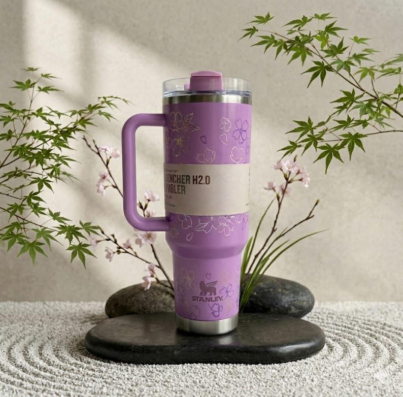 May include: A lavender Stanley Quencher H2.0 tumbler with a handle, adorned with a floral pattern. The tumbler has a clear lid and a silver base. The text "Quencher H2.0 Tumbler" is visible on a paper band around the middle. The Stanley logo is at the bottom.