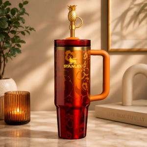 Pode incluir: Um copo Stanley vermelho e dourado com uma al&ccedil;a. O copo tem um topo em forma de abacaxi e um design floral. A al&ccedil;a e a borda s&atilde;o laranja. O logotipo da Stanley &eacute; dourado.