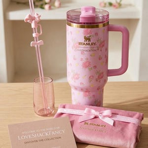 K&ouml;nnte beinhalten: Ein rosa Stanley Loveshackfancy Reisebecher mit floralem Muster, einem passenden Strohhalm, einem kleinen Glas, einem rosa Samtbeutel und einer Karte mit "Welcome to the World of Loveshackfancy".