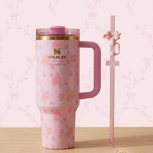 K&ouml;nnte beinhalten: Ein rosa Isolierbecher mit floralem Muster und passendem Griff. Der Becher hat einen goldenen Rand und einen rosa Deckel. Ein klarer Strohhalm mit rosa Schleifen ist enthalten. Der Text "STANLEY LOVESHACKFANCY" ist auf dem Becher aufgedruckt.