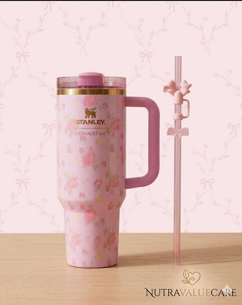 K&ouml;nnte beinhalten: Ein rosa Isolierbecher mit floralem Muster und passendem Griff. Der Becher hat einen goldenen Rand und einen rosa Deckel. Ein klarer Strohhalm mit rosa Schleifen ist enthalten. Der Text "STANLEY LOVESHACKFANCY" ist auf dem Becher aufgedruckt.