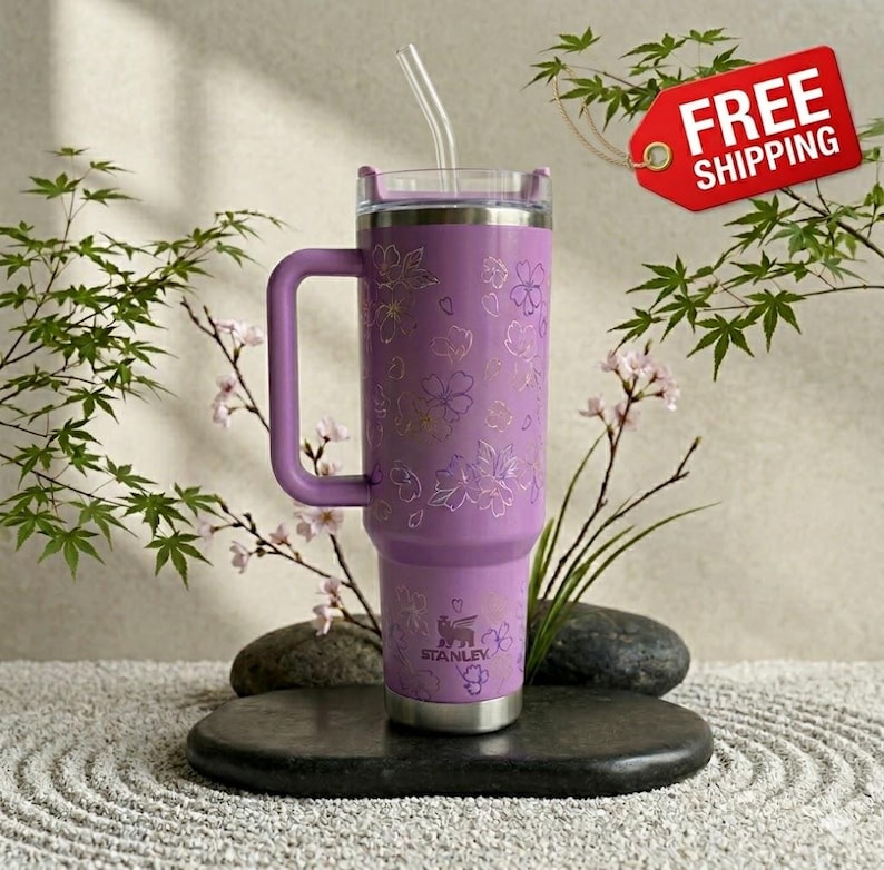 May include: A lavender Stanley tumbler with a handle and a clear straw. The tumbler features a floral design in gold and the Stanley logo. The tumbler is approximately 10 inches tall.