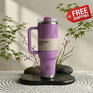 May include: A lavender Stanley Quencher H2.0 Flowstate tumbler with a handle, decorated with a floral pattern. The tumbler has a clear lid and a silver base. The text "Quencher H2.0 Tumbler" is visible on the side.