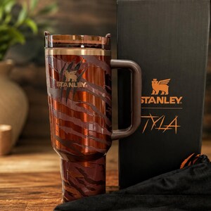 May include: A brown and dark orange tiger-striped Stanley tumbler with a handle and a translucent lid. The tumbler features the Stanley logo and is displayed next to a black box with the Stanley logo and the text Tyla printed in orange.