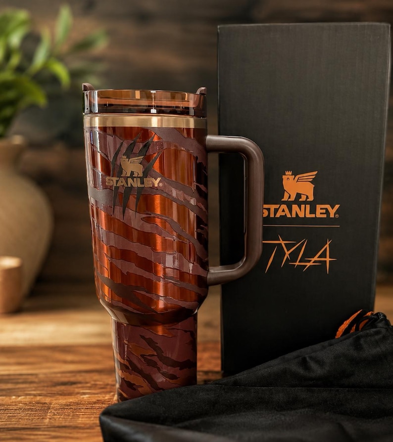 May include: A brown and dark orange tiger-striped Stanley tumbler with a handle and a translucent lid. The tumbler features the Stanley logo and is displayed next to a black box with the Stanley logo and the text Tyla printed in orange.