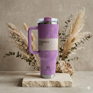 May include: A purple Stanley Quencher H2.0 tumbler with a handle, lid, and floral design. The tumbler is displayed on a stone block with dried plants in the background. The tumbler has a silver base.