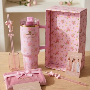 K&ouml;nnte beinhalten: Ein rosa Stanley-Becher mit Griff, einem durchsichtigen Strohhalm und einem rosa Glas, alle mit einem Blumenmuster verziert. Der Becher und die Box tragen den Text "LOVESHACKFANCY". Ein rosa Samtbeutel und eine Karte mit dem Text "WELCOME TO THE WORLD OF LOVESHACKFANCY" sind ebenfalls vorhanden.