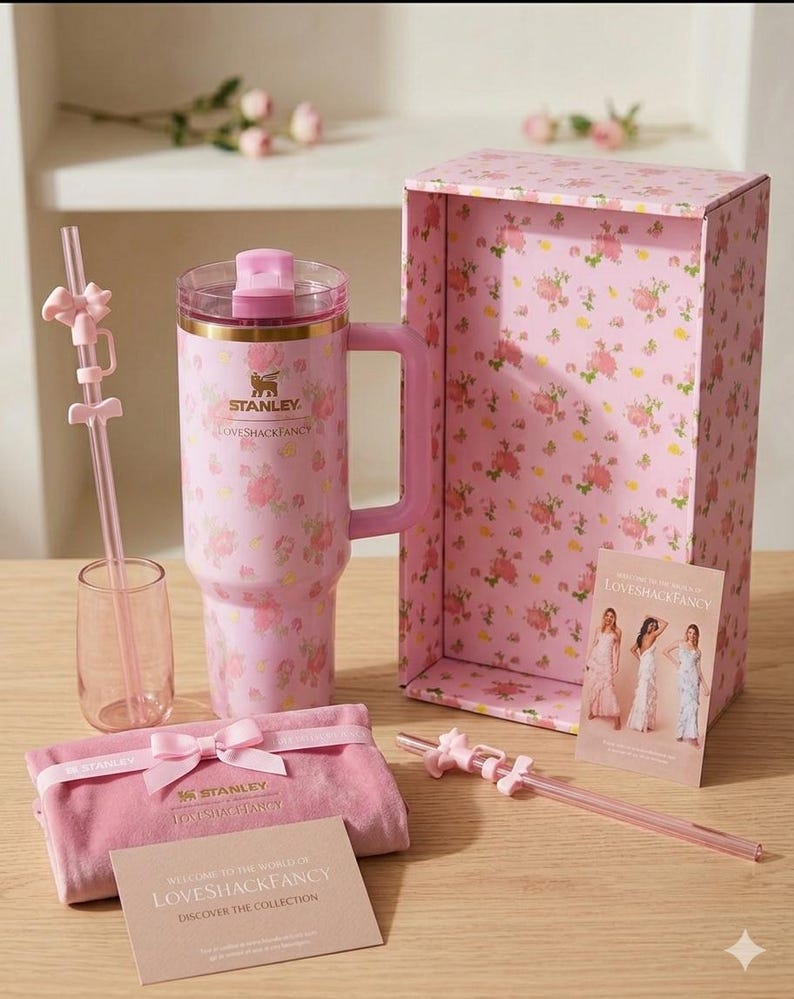 K&ouml;nnte beinhalten: Ein rosa Stanley-Becher mit Griff, einem durchsichtigen Strohhalm und einem rosa Glas, alle mit einem Blumenmuster verziert. Der Becher und die Box tragen den Text "LOVESHACKFANCY". Ein rosa Samtbeutel und eine Karte mit dem Text "WELCOME TO THE WORLD OF LOVESHACKFANCY" sind ebenfalls vorhanden.