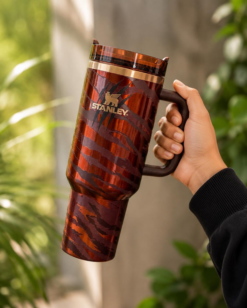 May include: A tall, cylindrical Stanley tumbler featuring a metallic copper and dark brown tiger stripe pattern. The drinkware includes a dark brown side handle, a transparent lid, and the gold Stanley logo printed on the upper front section.