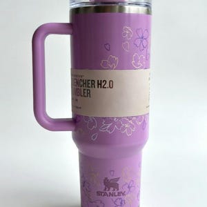 May include: A lavender Stanley Quencher H2.0 Flowstate Tumbler with a handle and floral design. The tumbler has a clear lid and a light pink accent on the top. The text "Quencher H2.0 Tumbler" is visible on the side.