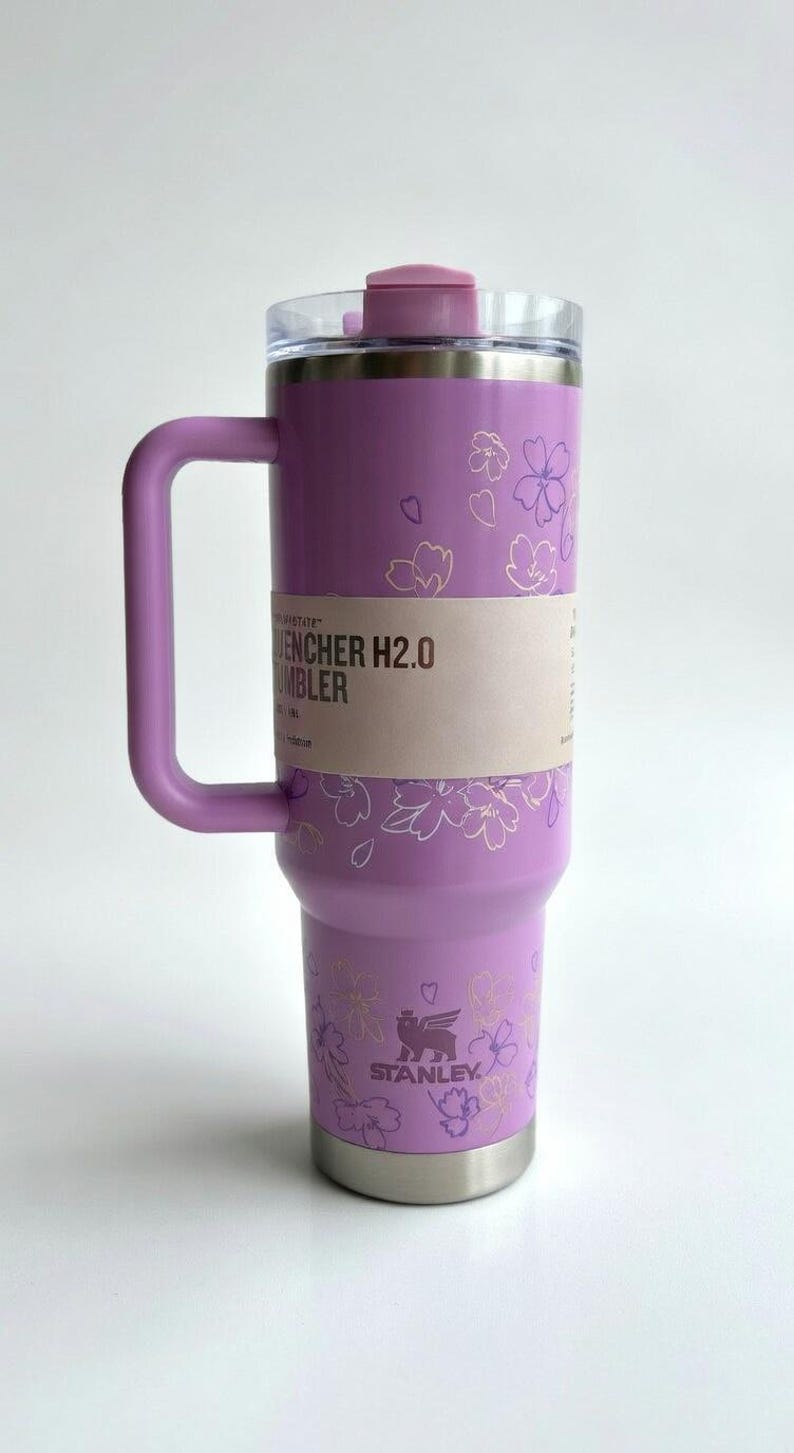May include: A lavender Stanley Quencher H2.0 Flowstate Tumbler with a handle and floral design. The tumbler has a clear lid and a light pink accent on the top. The text "Quencher H2.0 Tumbler" is visible on the side.