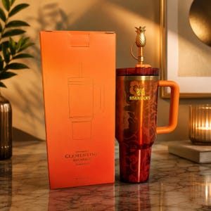 Pode incluir: Um copo Starbucks Clementine Quencher laranja com uma tampa em forma de abacaxi e uma al&ccedil;a a condizer. O copo tem um design floral e o log&oacute;tipo Starbucks. A caixa ao lado &eacute; laranja e tem um desenho do copo.