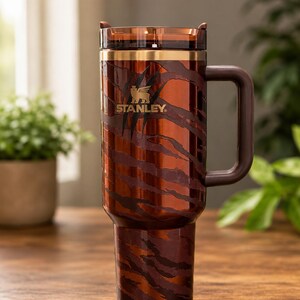 May include: A tall, cylindrical Stanley tumbler featuring a metallic copper and dark brown tiger stripe pattern. The cup includes a dark brown side handle, a gold-toned rim, and a translucent dark brown lid with a gold Stanley logo and claw marks.