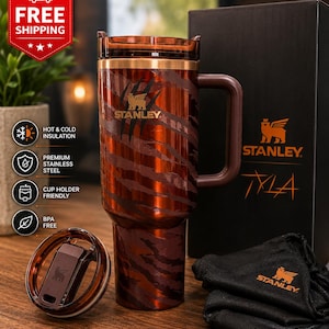 May include: A brown and copper-toned Stanley tumbler featuring a tiger-stripe pattern and claw mark graphics. The insulated cup includes a matching lid, a sturdy side handle, and is presented alongside a black branded box and a small black pouch.