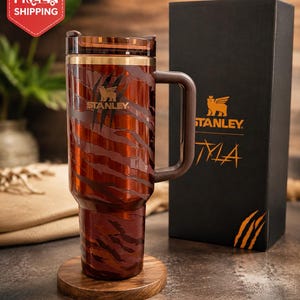 May include: A brown and orange tiger-striped Stanley tumbler with a handle, a clear lid, and a gold-tone rim. The cup features a Stanley logo and claw mark graphics. It is displayed next to a black box with the Stanley logo and the text TYLA.