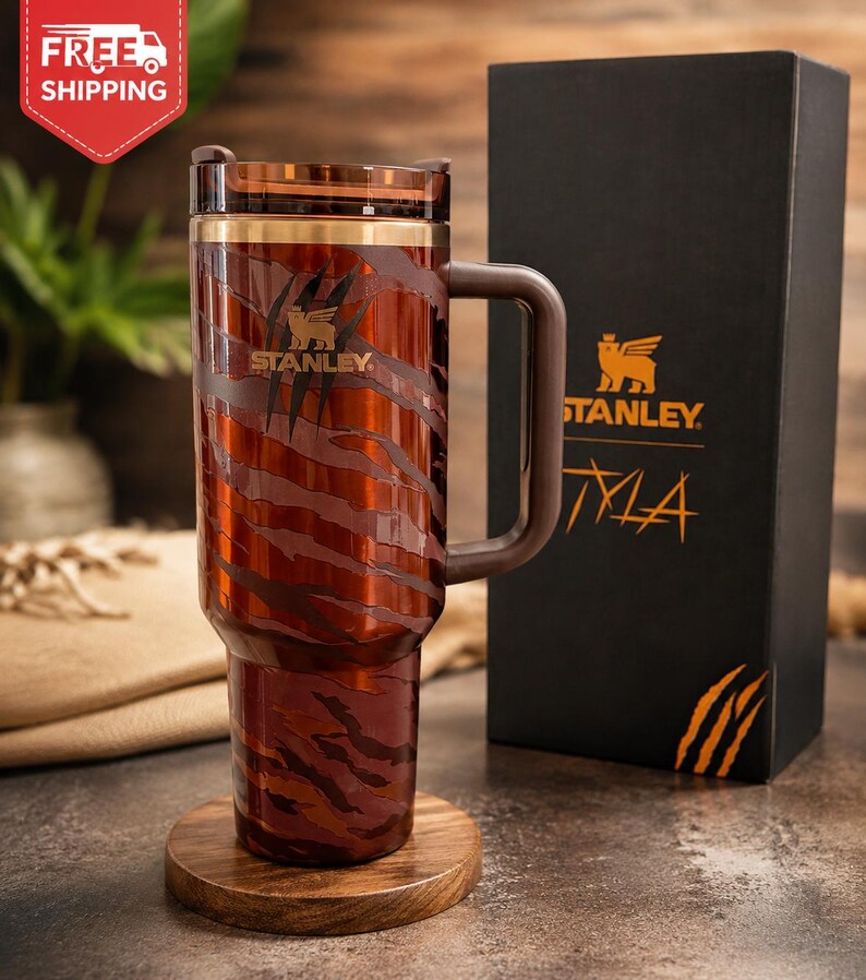 May include: A brown and orange tiger-striped Stanley tumbler with a handle, a clear lid, and a gold-tone rim. The cup features a Stanley logo and claw mark graphics. It is displayed next to a black box with the Stanley logo and the text TYLA.
