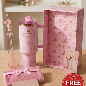 K&ouml;nnte beinhalten: Ein rosa Stanley-Becher mit Blumenmuster, Griff, passendem Strohhalm und kleinem Glas. Das Set enth&auml;lt eine rosa Schachtel mit Blumenmustern, einen Samtbeutel und eine Karte mit "LOVESHACKFANCY". Der Becher hat die Logos Stanley und LOVESHACKFANCY.