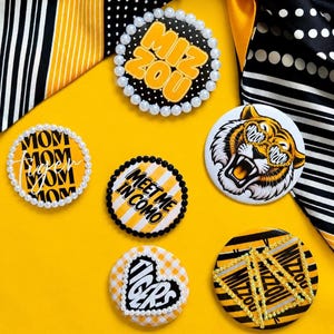 May include: Five circular pinback buttons featuring yellow, black, and white designs. One button says MIZ ZOU with pearls, one says MOM TIGER, one says MEET ME IN COMO, one features a tiger wearing MIZ ZOU glasses, and one has TIGERS text.