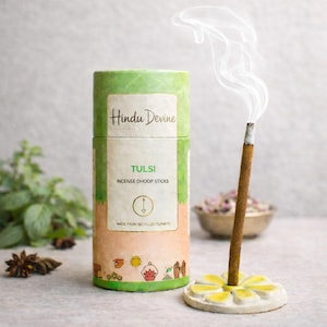 May include: A cylindrical container of Hindu Devine Tulsi incense sticks with a green and beige design. A lit incense stick emits a stream of white smoke, placed in a white and yellow flower-shaped holder. The text "TULSI INCENSE DHOOP STICKS" is visible.